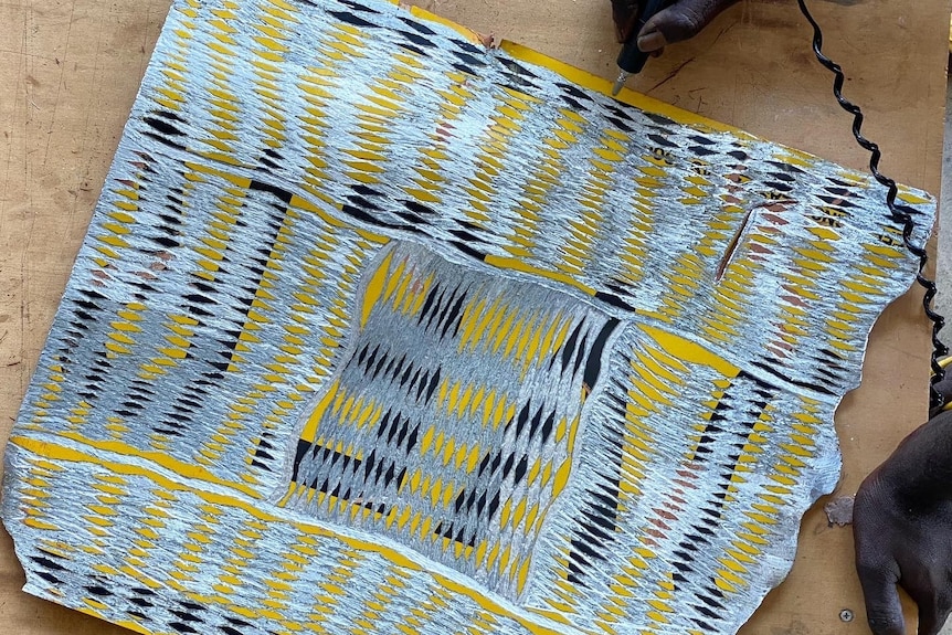 A hand can be seen etching a patterned design onto an old road sign in silver, black and yellow