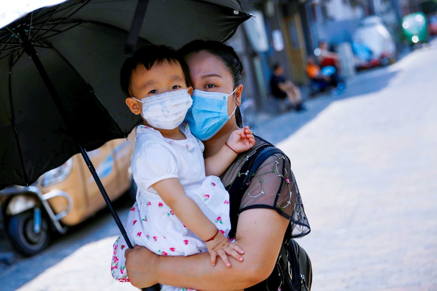 A woman in a blue face masking holding a baby in a floral mask under an umbrella