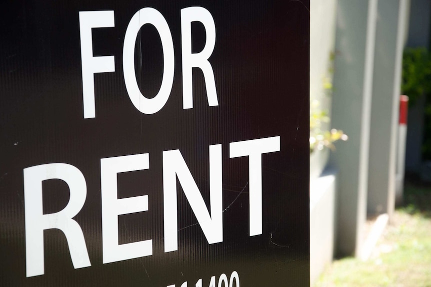 A black and white 'For Rent' sign displayed outside a block of apartments.