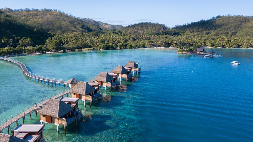 A line of huts sit on stilts in clear blue water with a white beach and mountains covered in trees in the background.