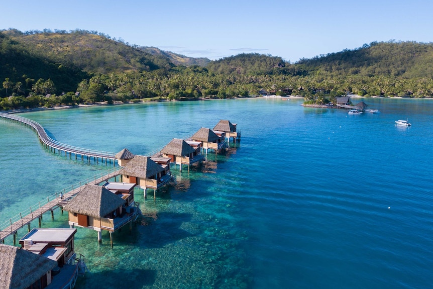 A line of huts sit on stilts in clear blue water with a white beach and mountains covered in trees in the background.
