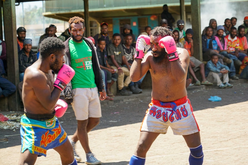 The men face each other wearing gloves and shin guards, as an official watches on.