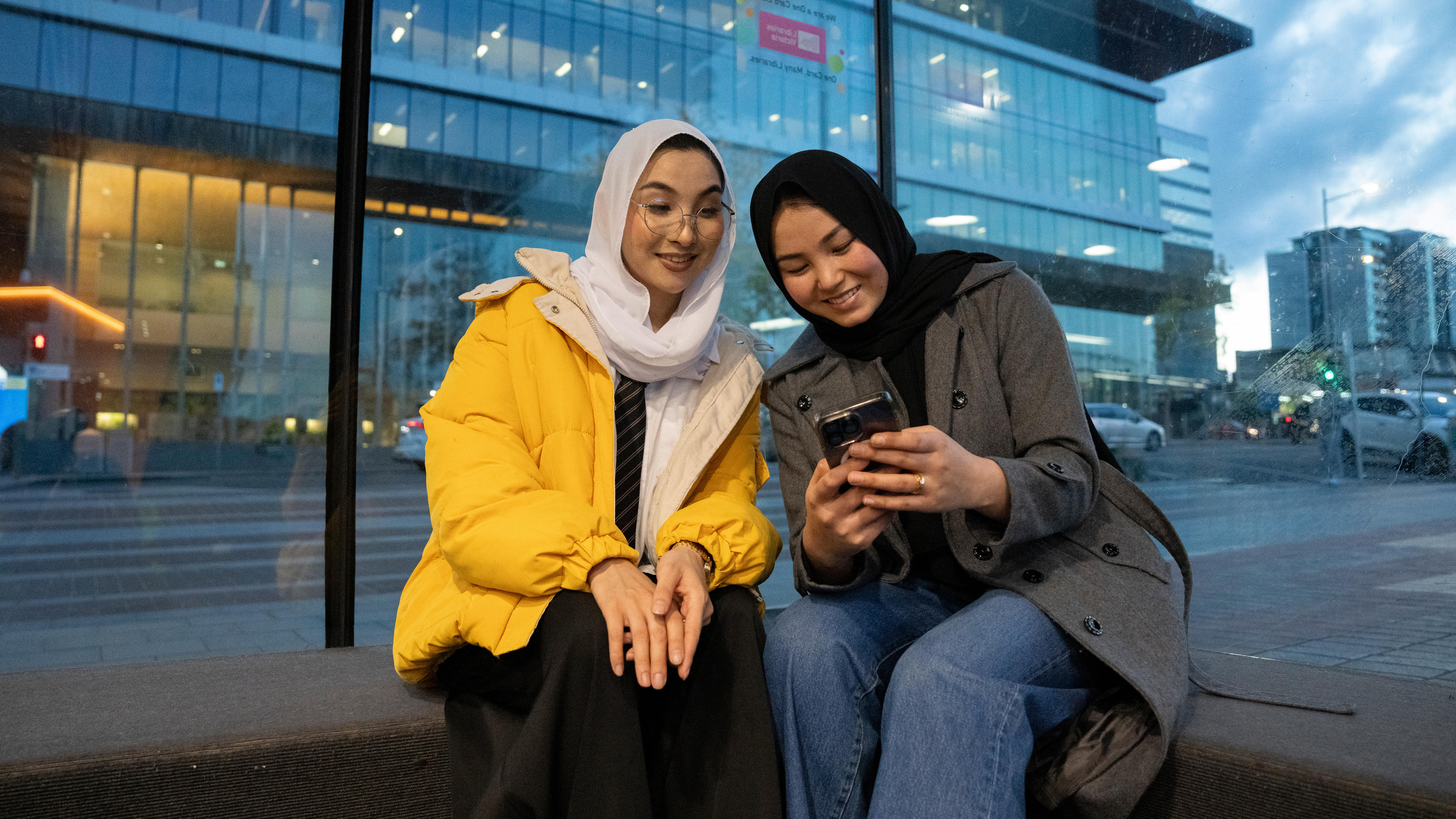 Two sisters, in a yellow and grey coat, sit on the phone video conferencing in front of a window