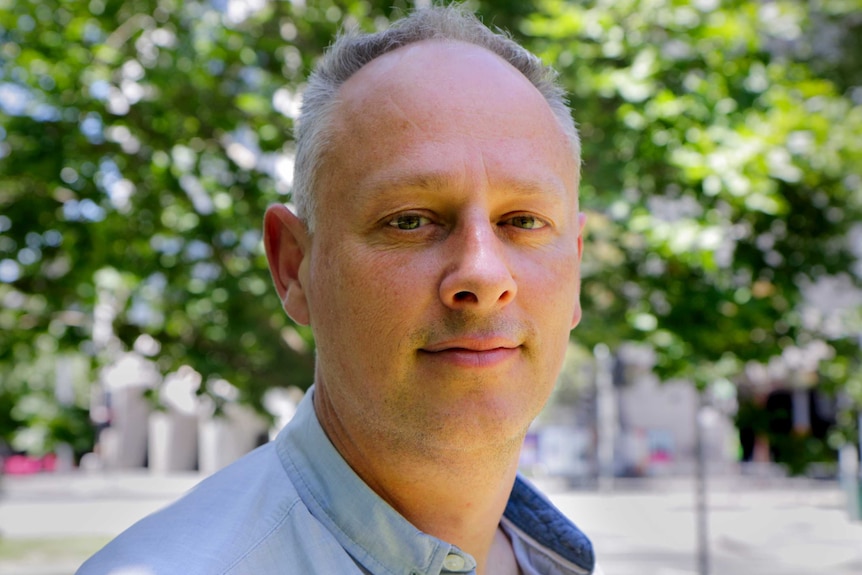 Man with grey hair and blue shirt smiles with green leaves of a tree behind him.