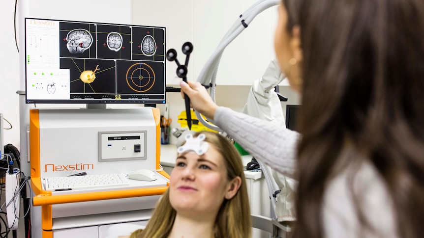 A young woman sits back in a chair while a doctor holds a scanner to her scalp. Brain images are on a monitor in the background