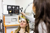A young woman sits back in a chair while a doctor holds a scanner to her scalp. Brain images are on a monitor in the background