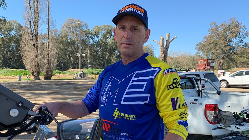 Man looks straight at camera, standing behind a motor bike and wearing racing gear.