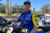 Man looks straight at camera, standing behind a motor bike and wearing racing gear.