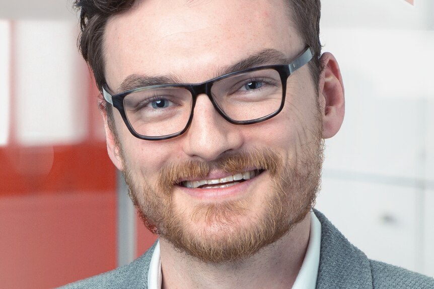 Headshot smiling man, brown hair, ginger beard, wears blue suit jacket, white shirt, black-rimmed glasses, background blurred.