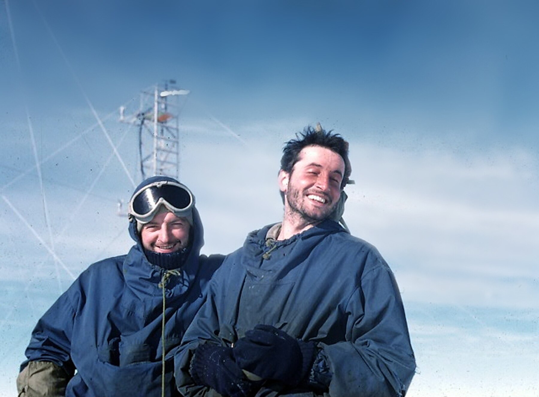 Two men stand in snowy conditions in front of a radio tower.