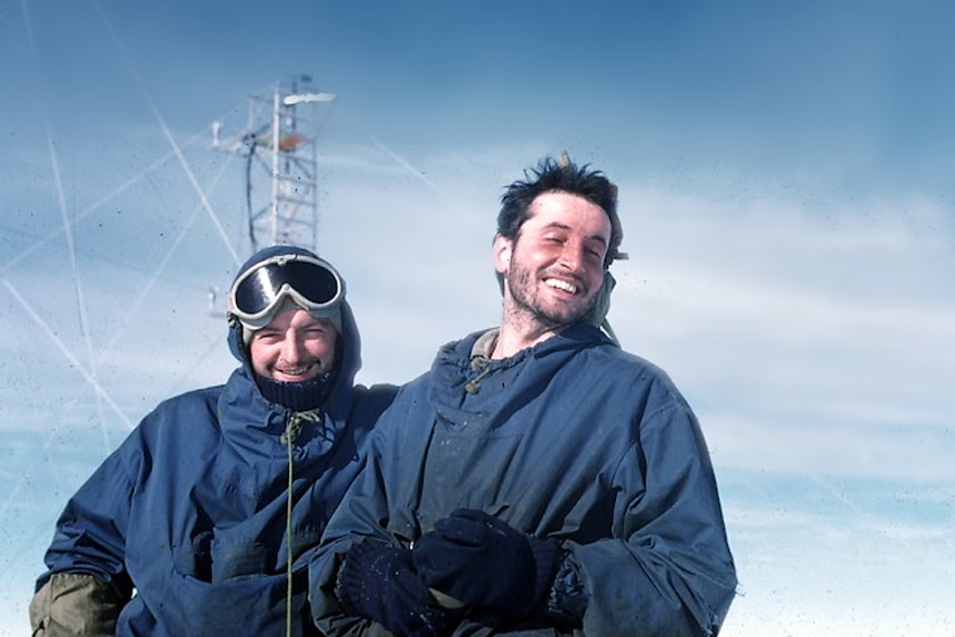 Two men stand in snowy conditions in front of a radio tower.
