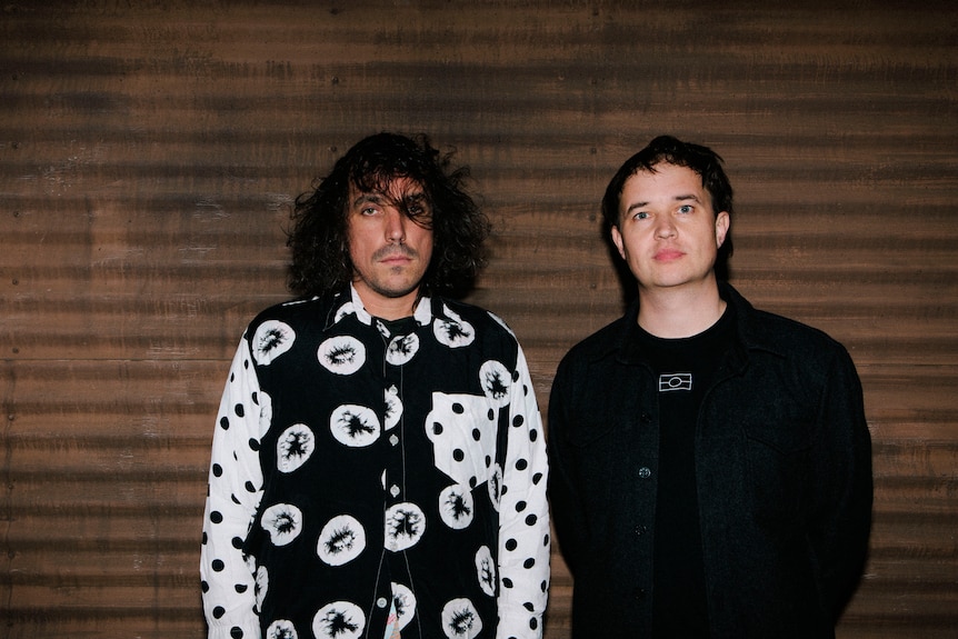 A man wearing a white and black shirt with long hair, and another man in a black top.