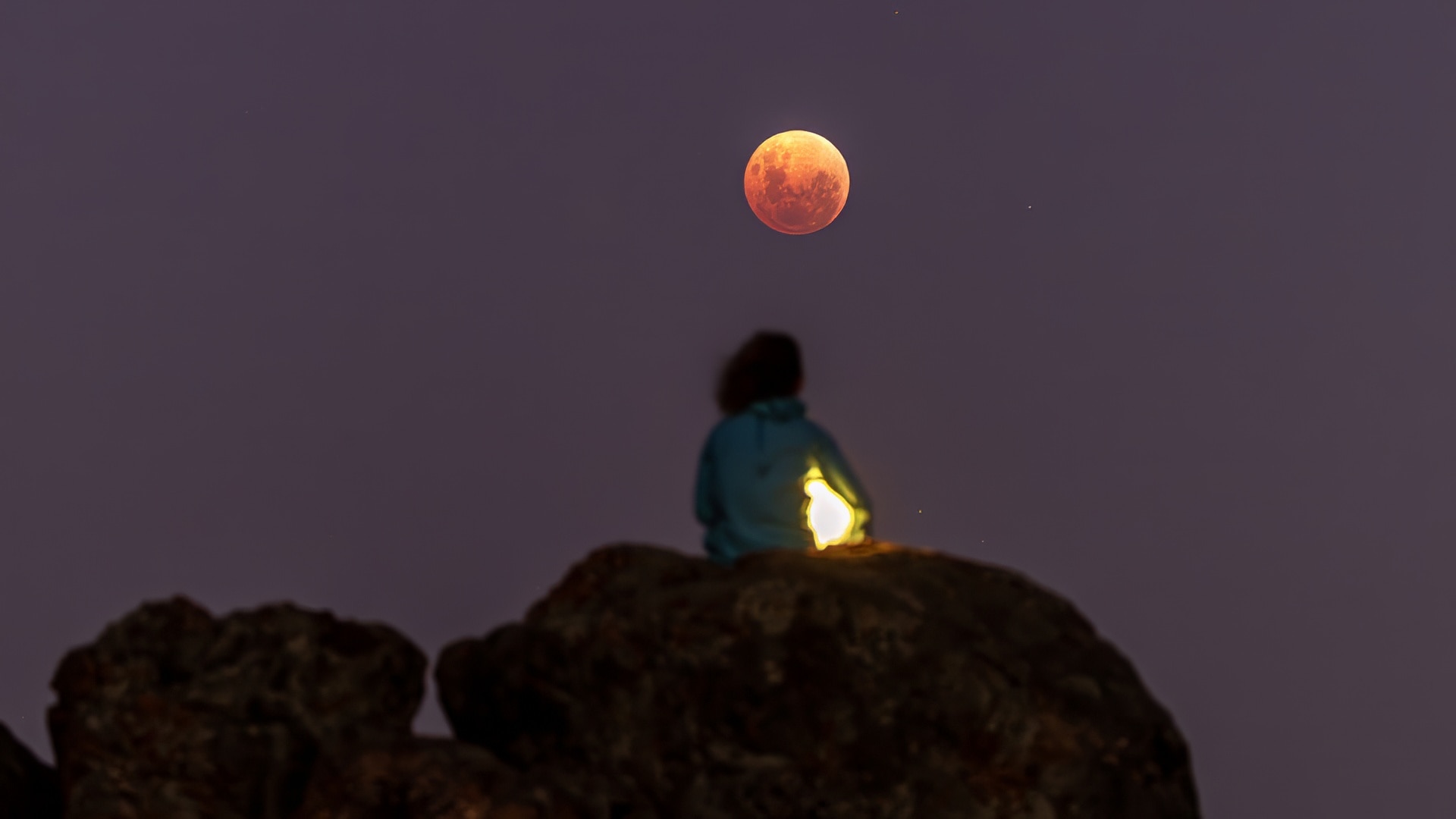 A woman sits on rocks looking at the blood moon.