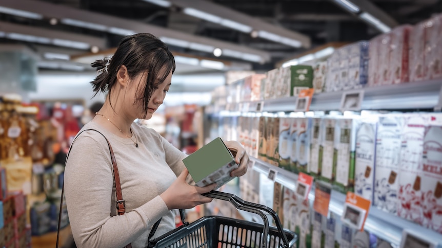 A picture of a young Asian woman looking at the label of a back of a product box in the supermarket aisle.