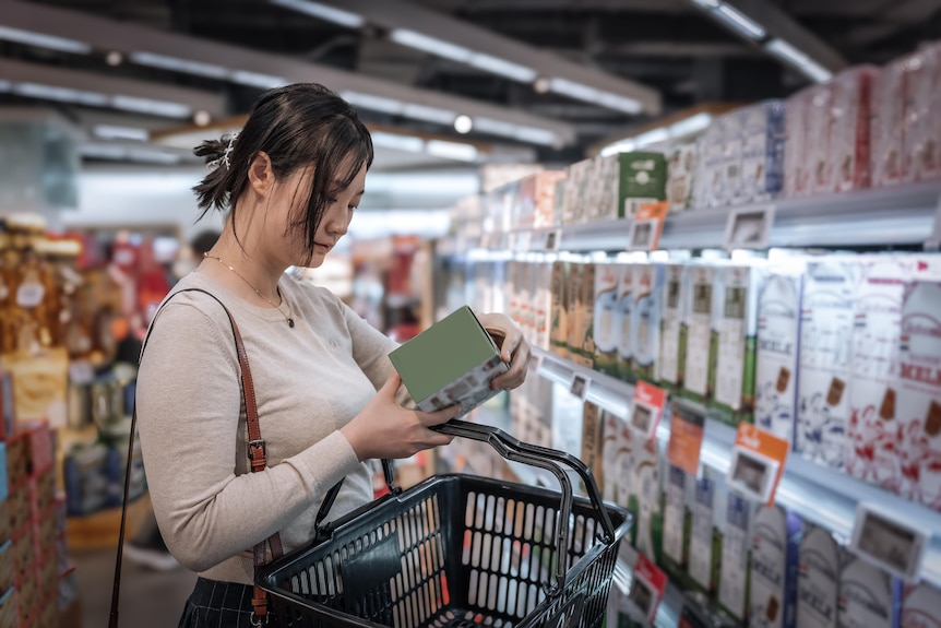 A picture of a young Asian woman looking at the label of a back of a product box in the supermarket aisle.