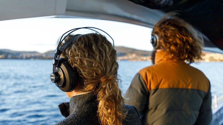 The back of a man and woman's heads with headphones on, standing on a boat, looking out at the water