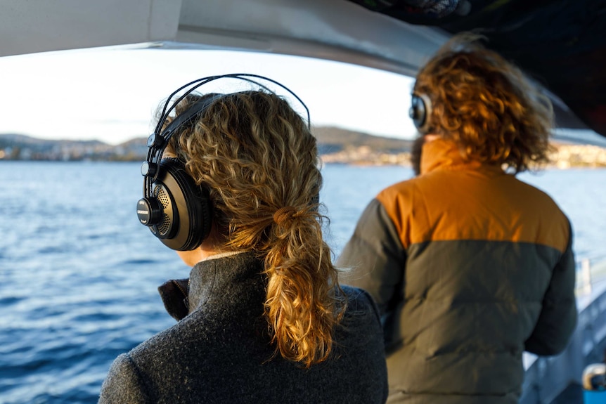 The back of a man and woman's heads with headphones on, standing on a boat, looking out at the water