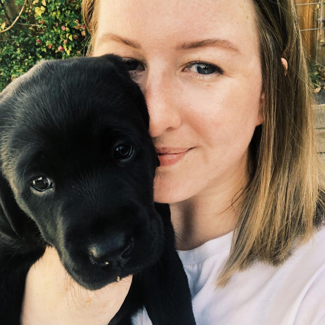 A blonde white woman in her 30s holds a black labrador puppy up to her face for a selfie 