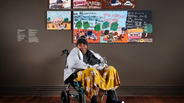 A woman in a wheelchair sits in a gallery in front of several paintings.