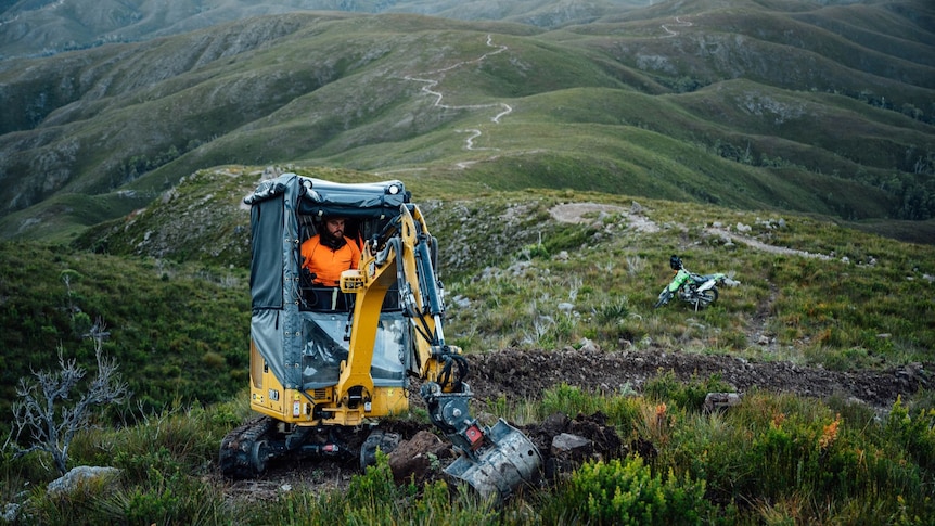 Small digger operating on remote mountain ridge lines, motorbike in the background.