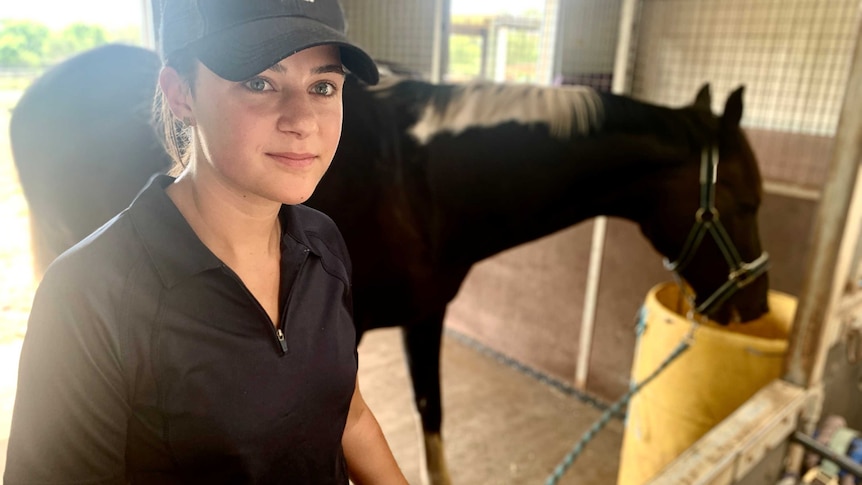 Maddie Nelson stands in a stable wearing a baseball cap, holding her horse, Pablo Cruize, by a rope