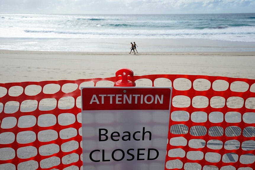 a closed beach sign with people walking next to the shoreline in the background