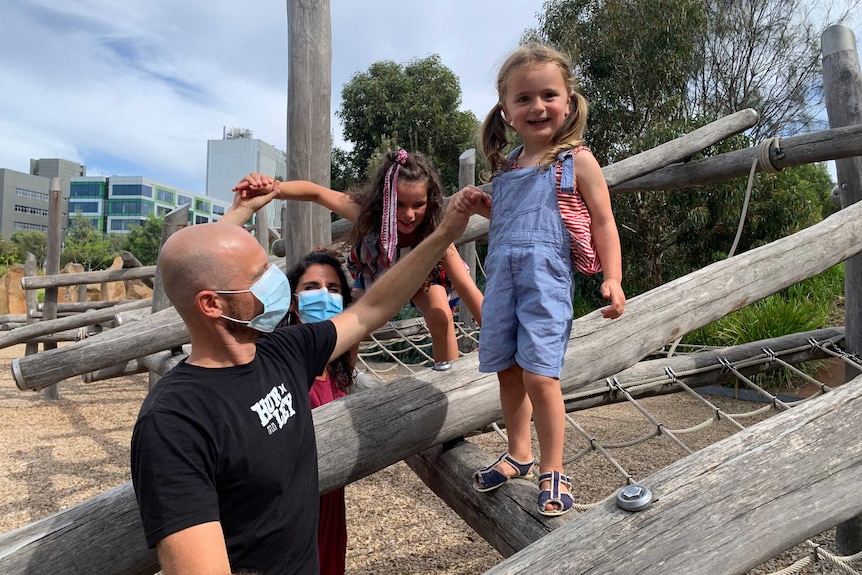 Francesca and Alessandro stand on the ground while holding their childrens' hands as they stand on play equipment.