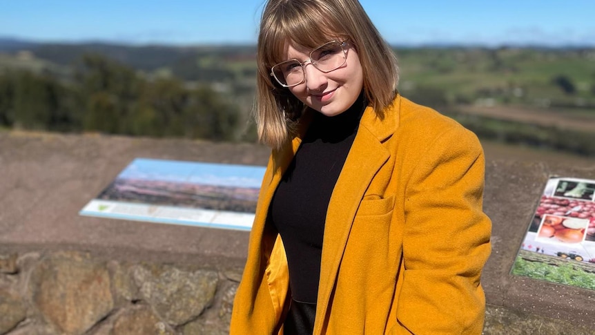 A young woman wearing a yellow coat and glasses smiles at the camera.