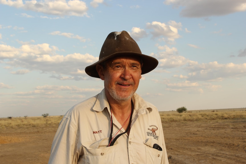Man in a hat wearing a beige shirt with sky and plains behind him