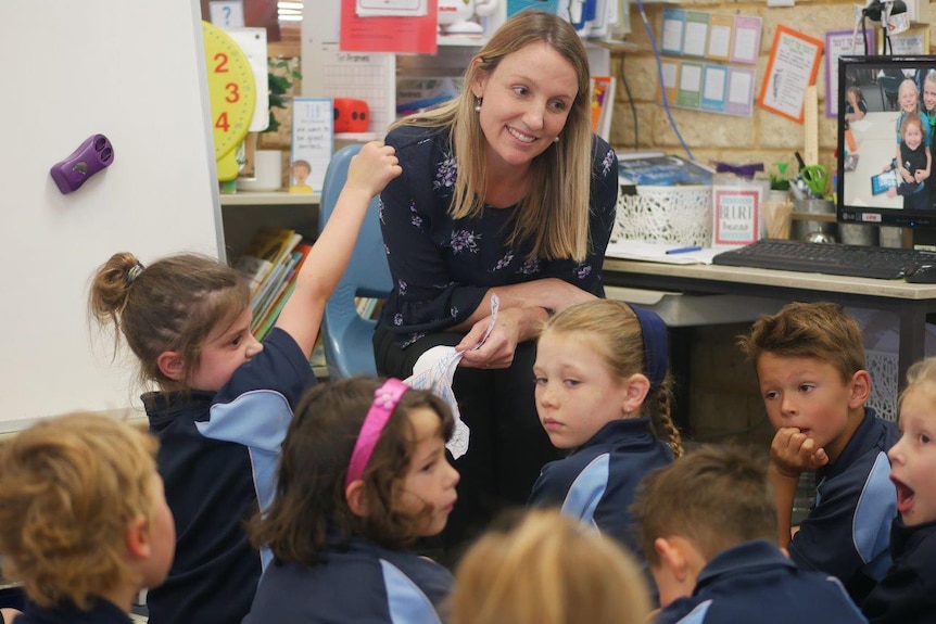 A smiling female teacher sits in front of a group of students sitting on the floor in class.