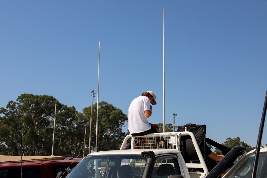 A young man wearing a white t shirt and cap sits on the back frame of a ute behind two goal posts 