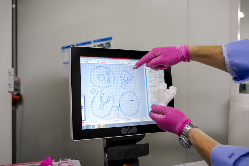 Man with blue sleeves  holds up small set of white tubes next to computer screen wearing pink gloves in a lab type setting