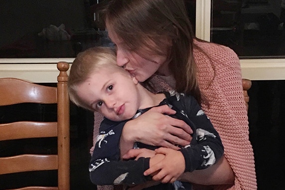 Woman kisses her son with blonde hair picture with cake with dinosaur figures.