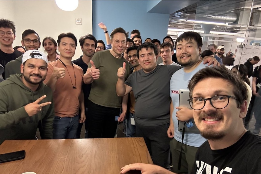 Elon Musk in a boardroom with a group of men, all giving thumbs up or peace signs