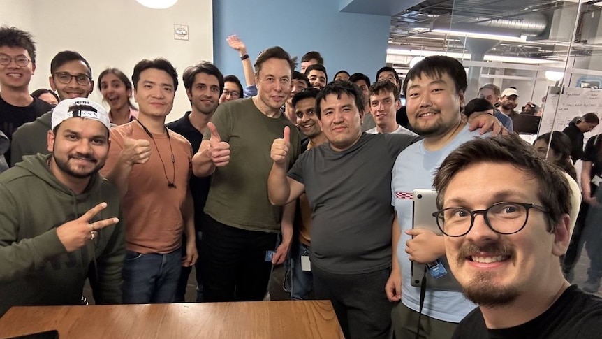 Elon Musk in a boardroom with a group of men, all giving thumbs up or peace signs