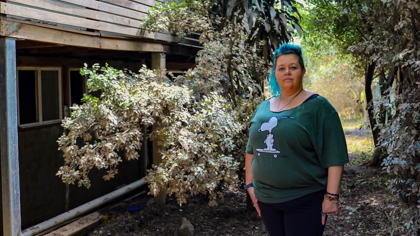 An image of Terri standing in front of trees/shrubs which are caked in mud and side of house 