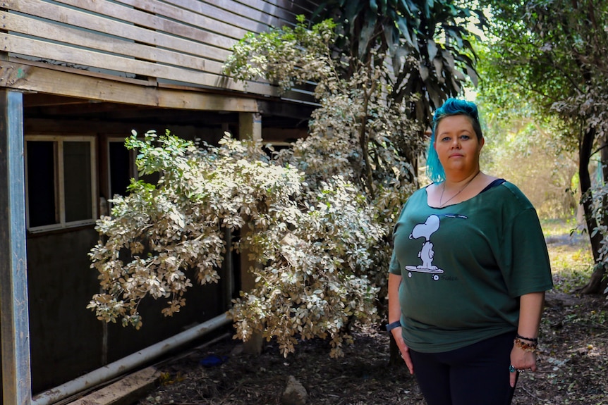 An image of Terri standing in front of trees/shrubs which are caked in mud and side of house 