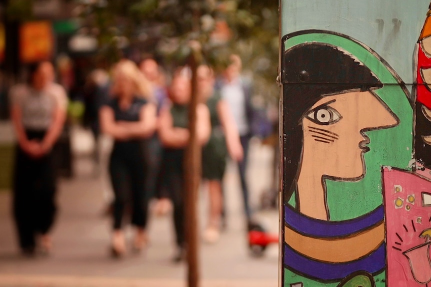 A painted electrical box in the foreground with people walking in the background.