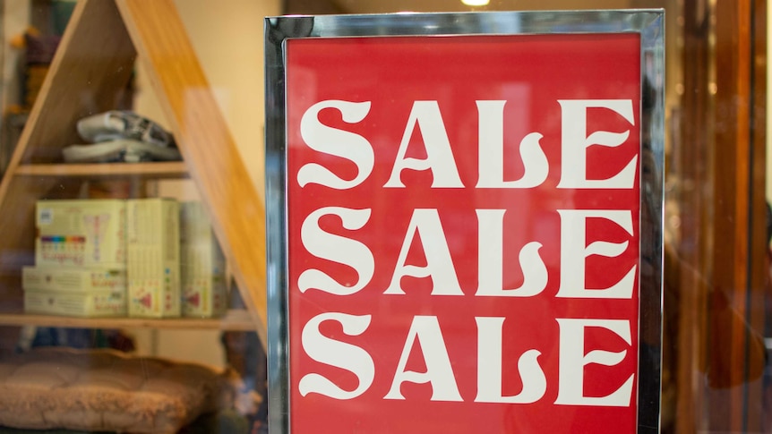 Sign says 'SALE SALE SALE' in a Brisbane shop window with merchandise in the background.