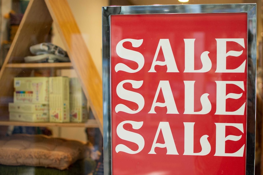 Sign says 'SALE SALE SALE' in a Brisbane shop window with merchandise in the background.