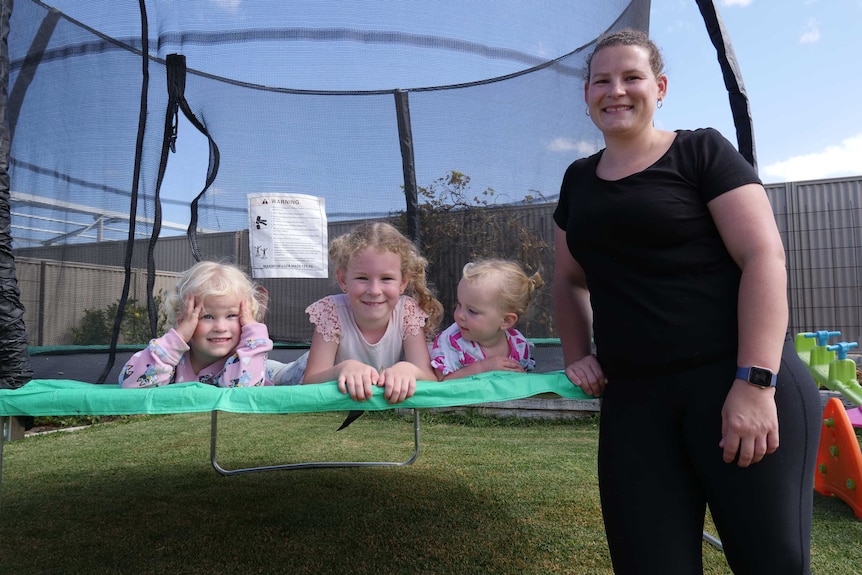 Three small children lie on a trampoline side-by-side while their mum stands near them