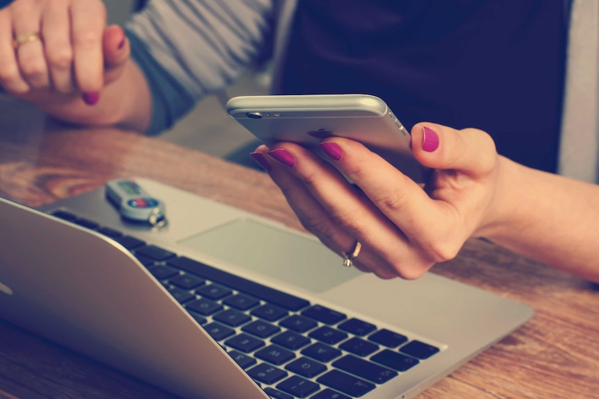 A woman using a laptop looks at her phone.