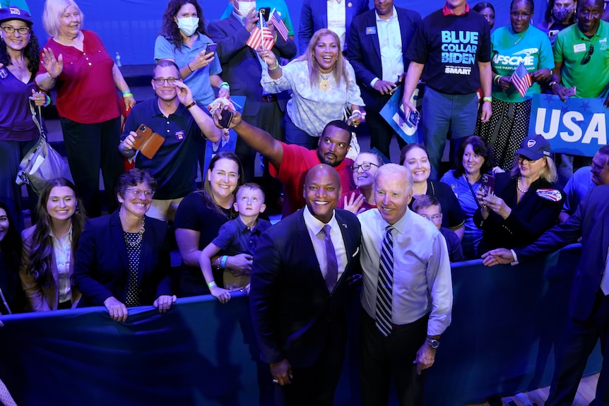 Two men with their arms around each other smile at the camera as people behind them also smile. 