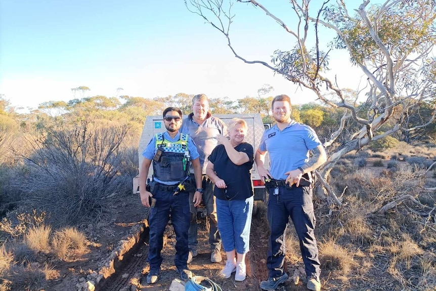 A smiling, middle-aged man and woman and two policemen stand in front of a vehicle on a bush track.