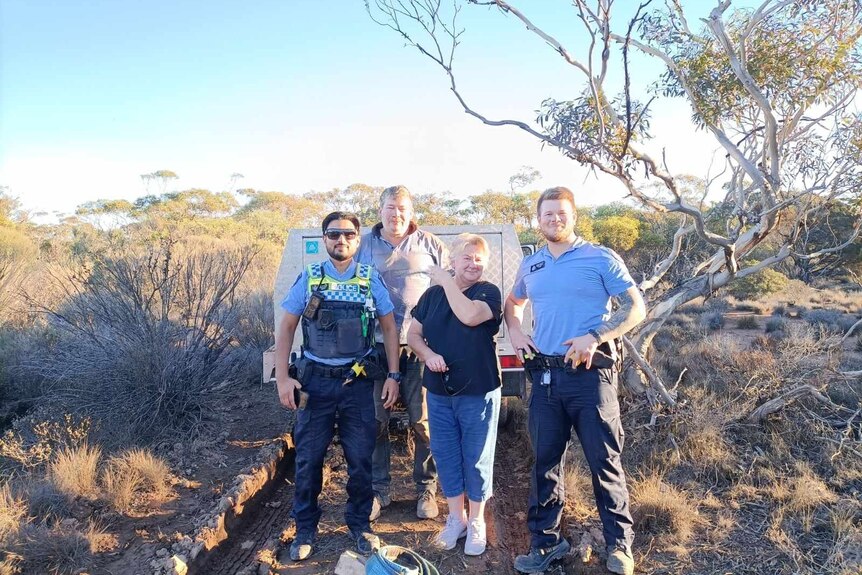 A smiling, middle-aged man and woman and two policemen stand in front of a vehicle on a bush track.