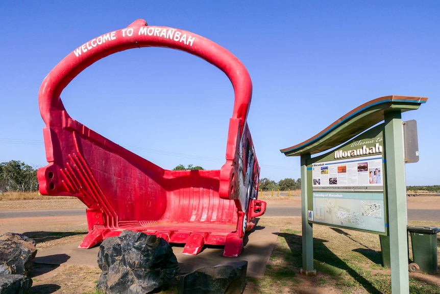 A large red dragline bucket next to a dusty sign.