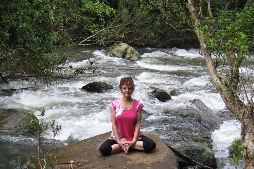A woman sits with her legs crossed in a yoga pose, on a rock amid rushing river rapids and surrounding bush.