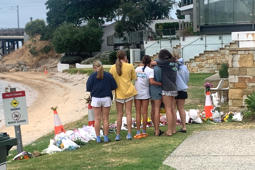 Six girls stand on the shore of the Swan River consoling one another, with their backs to the camera and flowers on the ground.