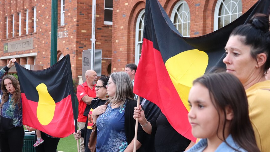 Women stand in front of red brick buildings, holding Aboriginal flags