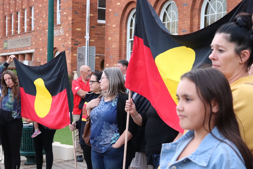 Women stand in front of red brick buildings, holding Aboriginal flags